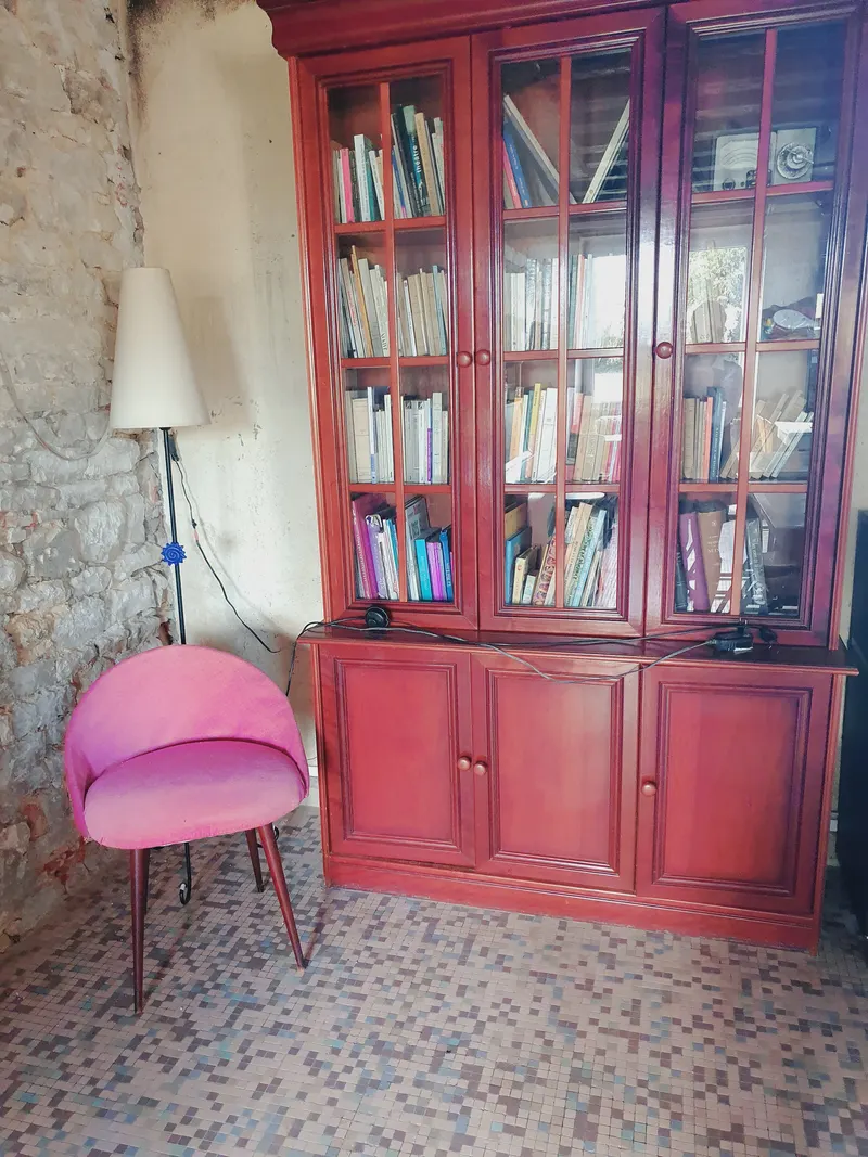 Une bibliothèque rouge vitrée remplie de livres, un fauteuil rose devant.