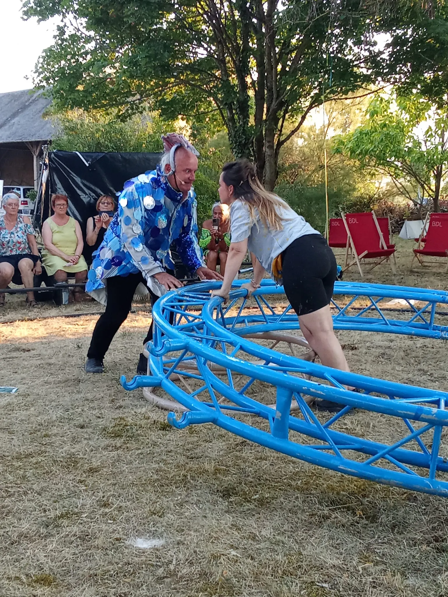 Dans la cour, sous les arbres, des artistes font rouler un grand cercle bleu devant le public.