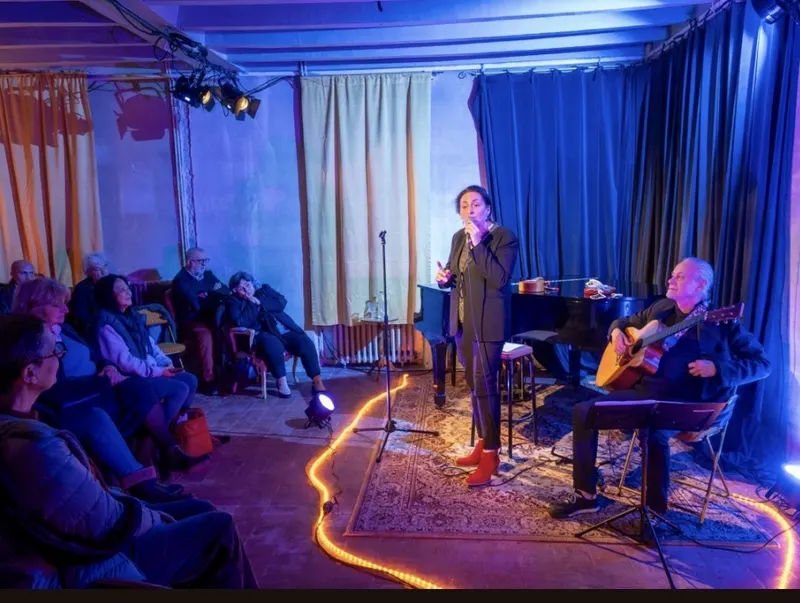 Une chanteuse et un guitariste devant un rideau bleu, le public assis tout près.