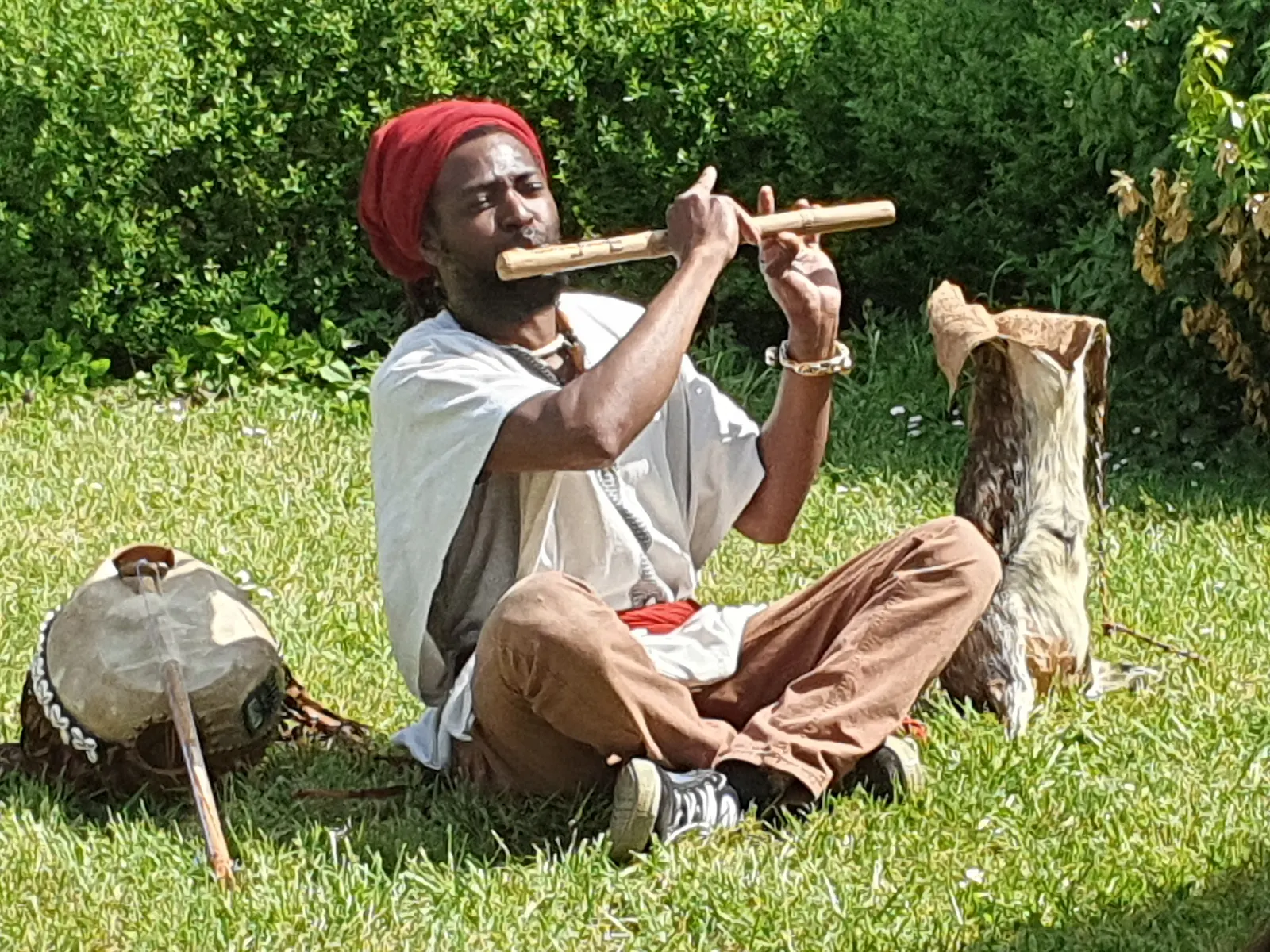 Yomba, assis dans l'herbe au soleil, joue d'une flûte en bois à côté de ses instruments.